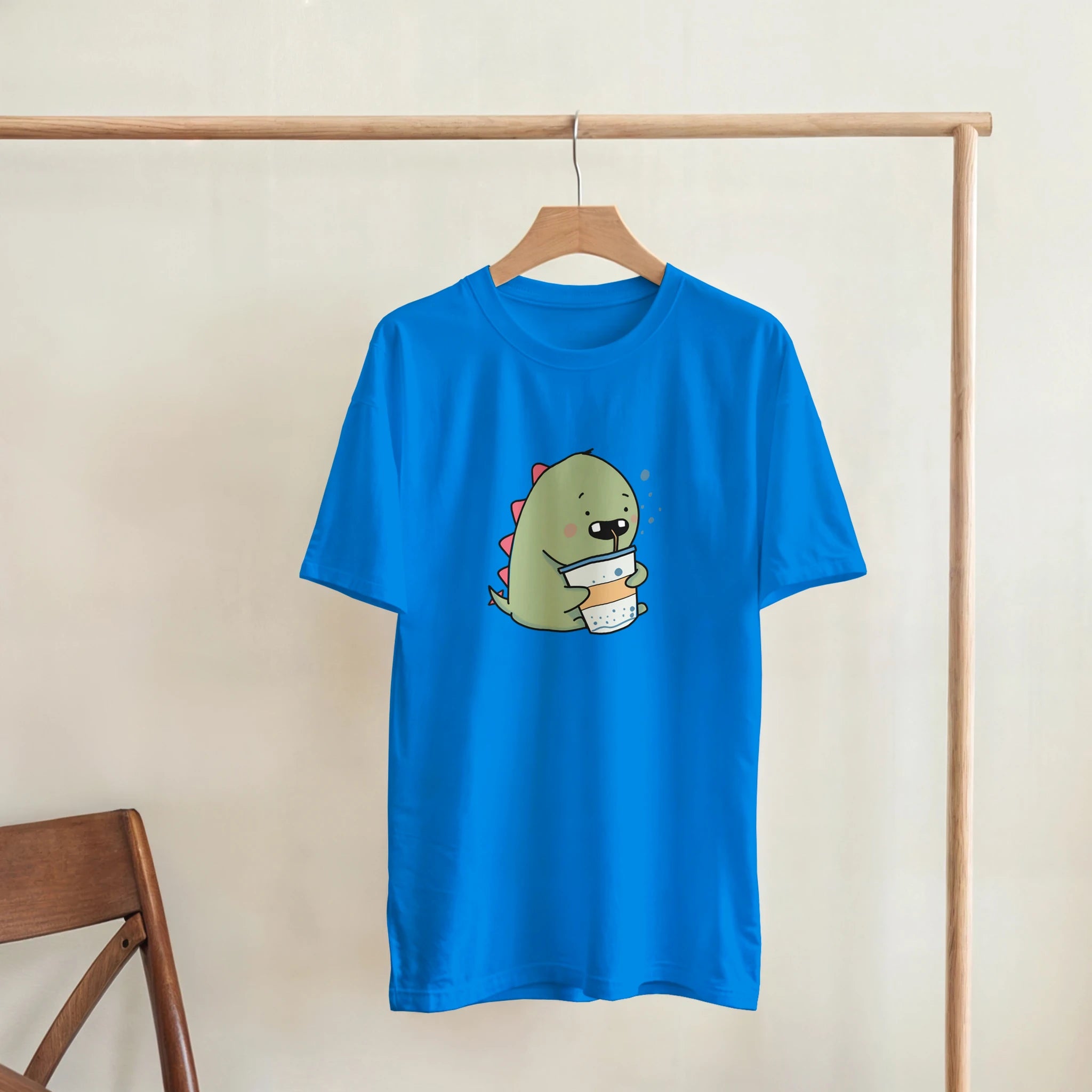A blue t-shirt with a cartoon dinosaur holding a cup of coffee, hanging on a wooden hanger.