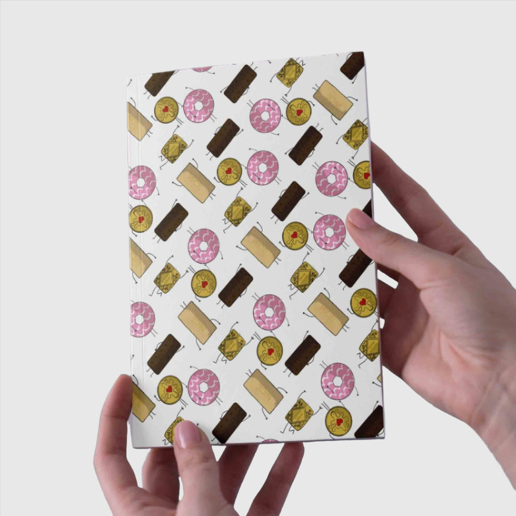 A hand holding an A5 notebook with a whimsical biscuit pattern cover featuring illustrated chocolate bars, jammy dodgers, custard creams, and lemon slices on a white background.”