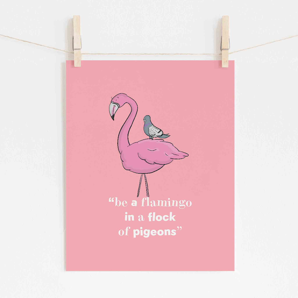 A playful pink flamingo illustration with a small pigeon perched on its back, set against a pastel pink background. Below the flamingo, a whimsical quote in a casual script reads 'be a flamingo in a flock of pigeons', all presented as a hanging wall art print.