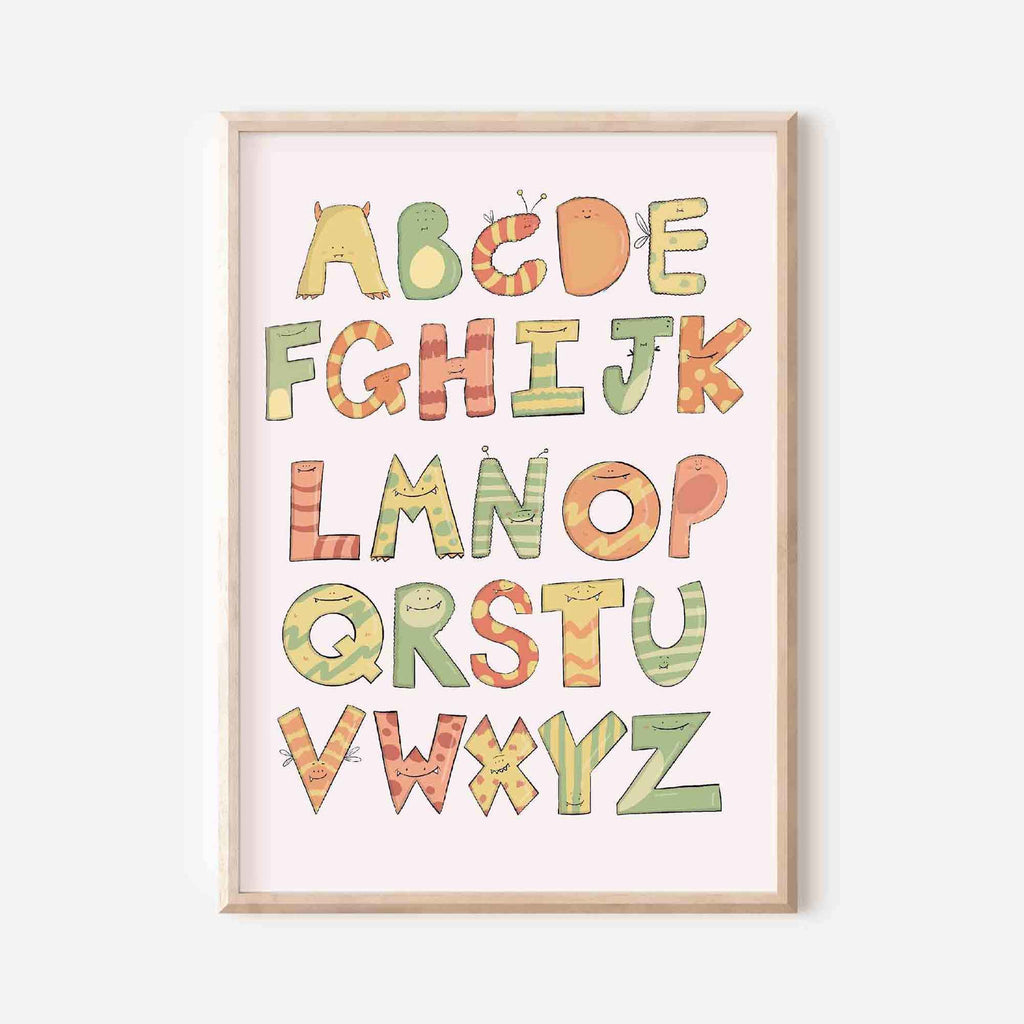 Colourful alphabet art print with each letter designed as a different playful monster character, presented in a variety of pastel tones against a light pink background, encased in a simple wooden frame.