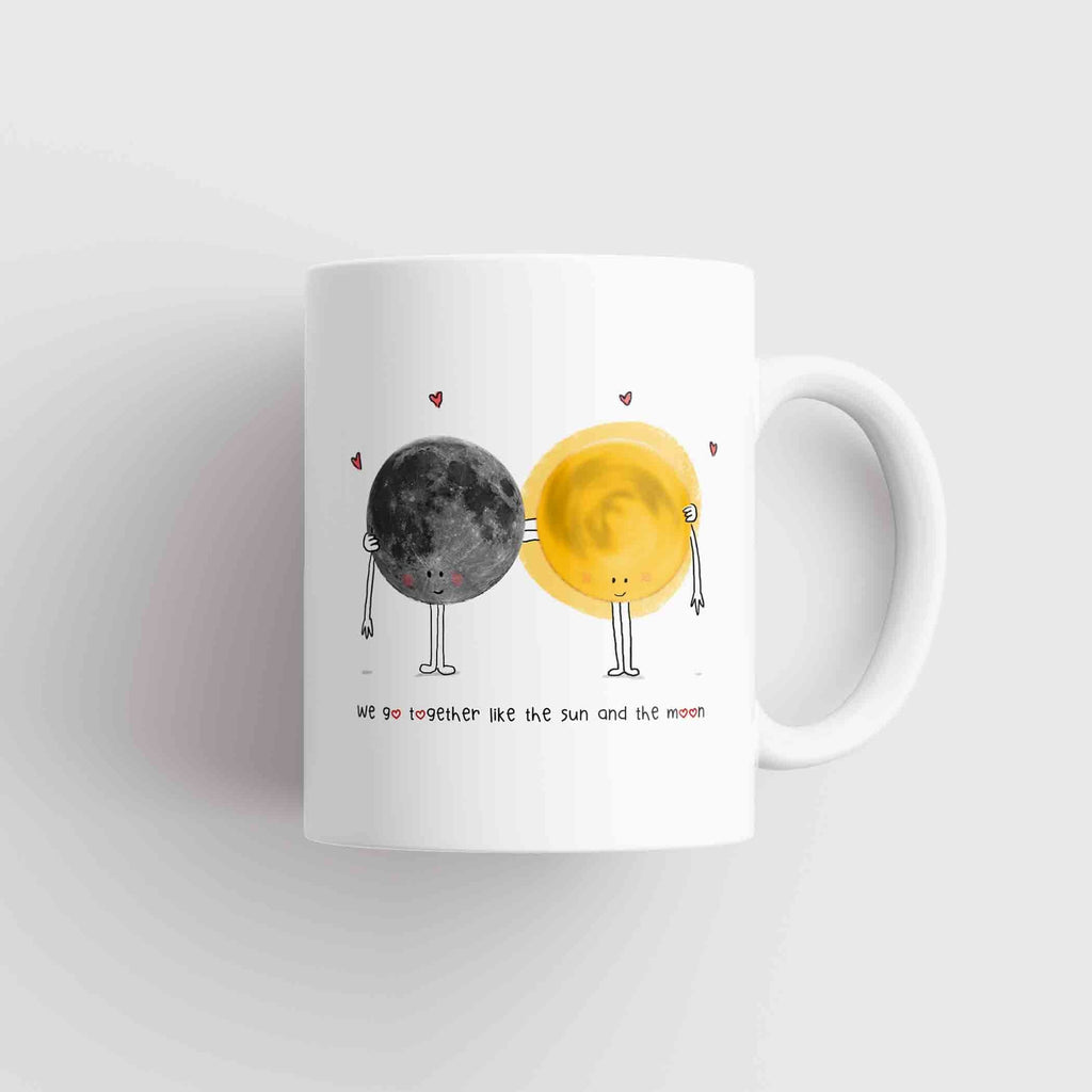 A white coffee mug with an artistic representation of the sun and moon with cute faces and arms, the sun in yellow and the moon in gray, with small red hearts floating above them. The phrase "we go together like the sun and the moon" is written in a playful, casual font below the illustration.